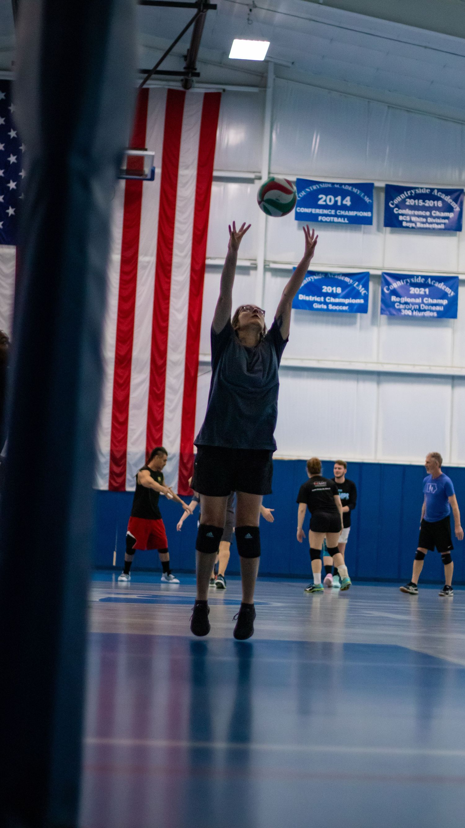 Player jumping to set a volleyball in a blue-floored indoor court with American flag on wall.