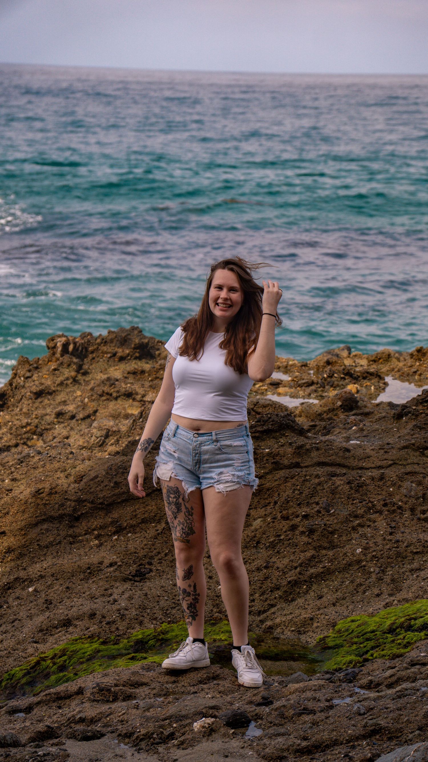 Individual wearing white crop top and shorts poses on coastal volcanic rocks.