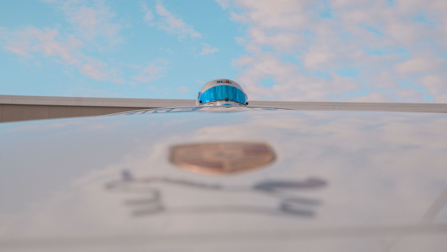Porsche badge on white car hood with blue helmet reflection against pastel sky.