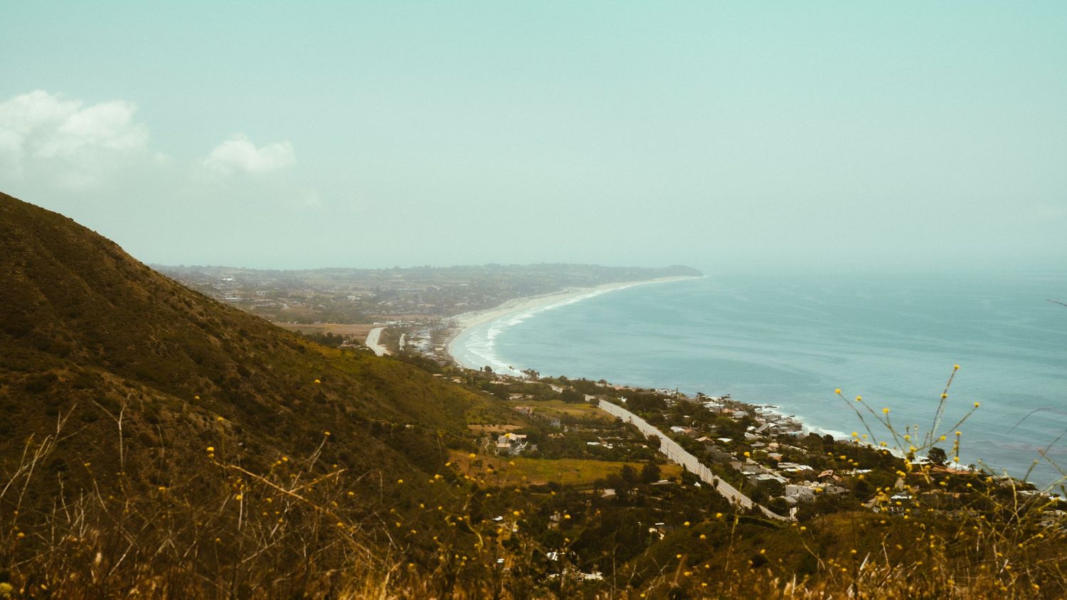 Panoramic view of a coastal town along a curved bay with hills and turquoise waters.