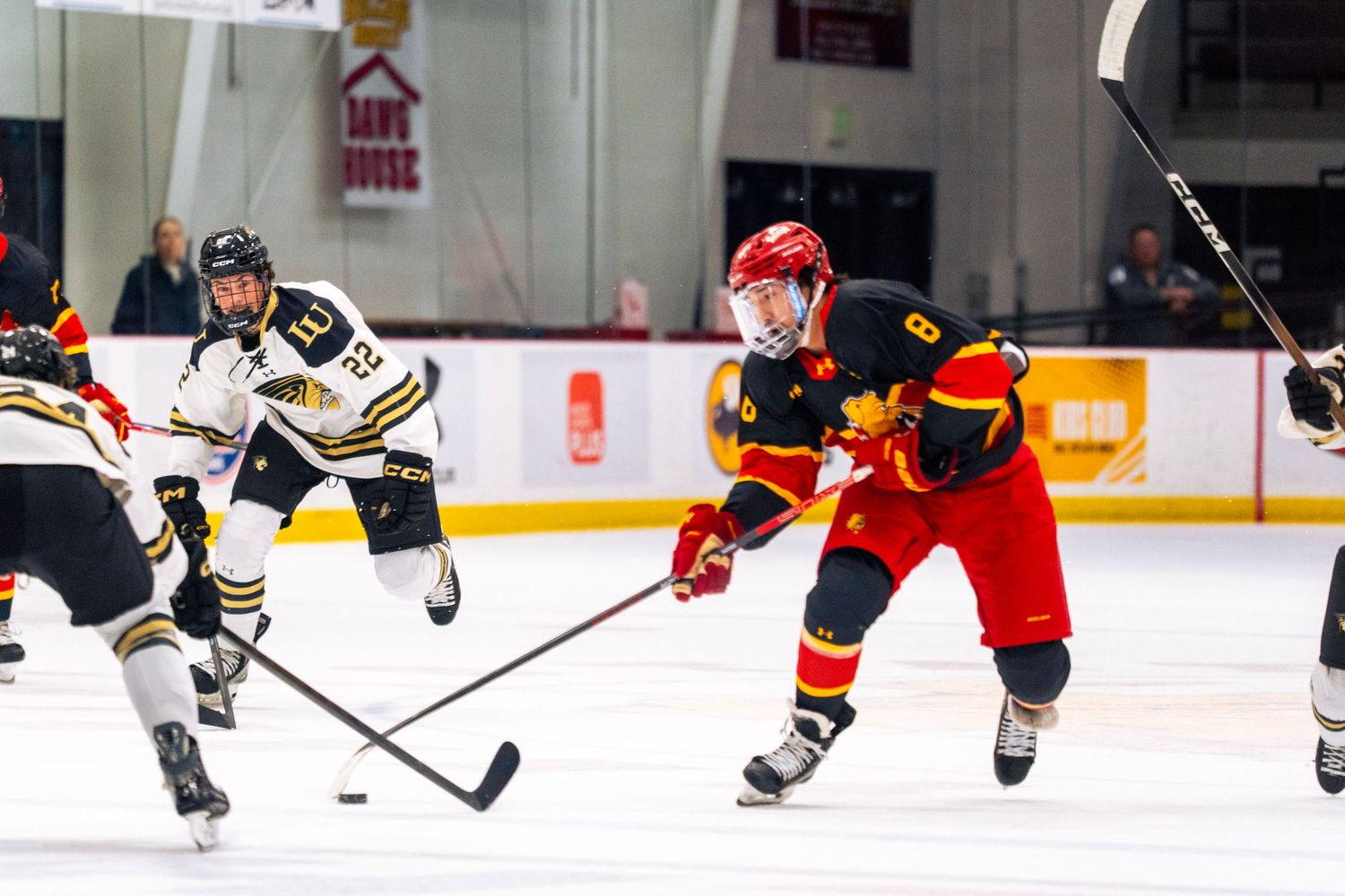 College hockey player in a red and gold jersey skates with the puck on the ice while defenders in white and black close in during a game.
