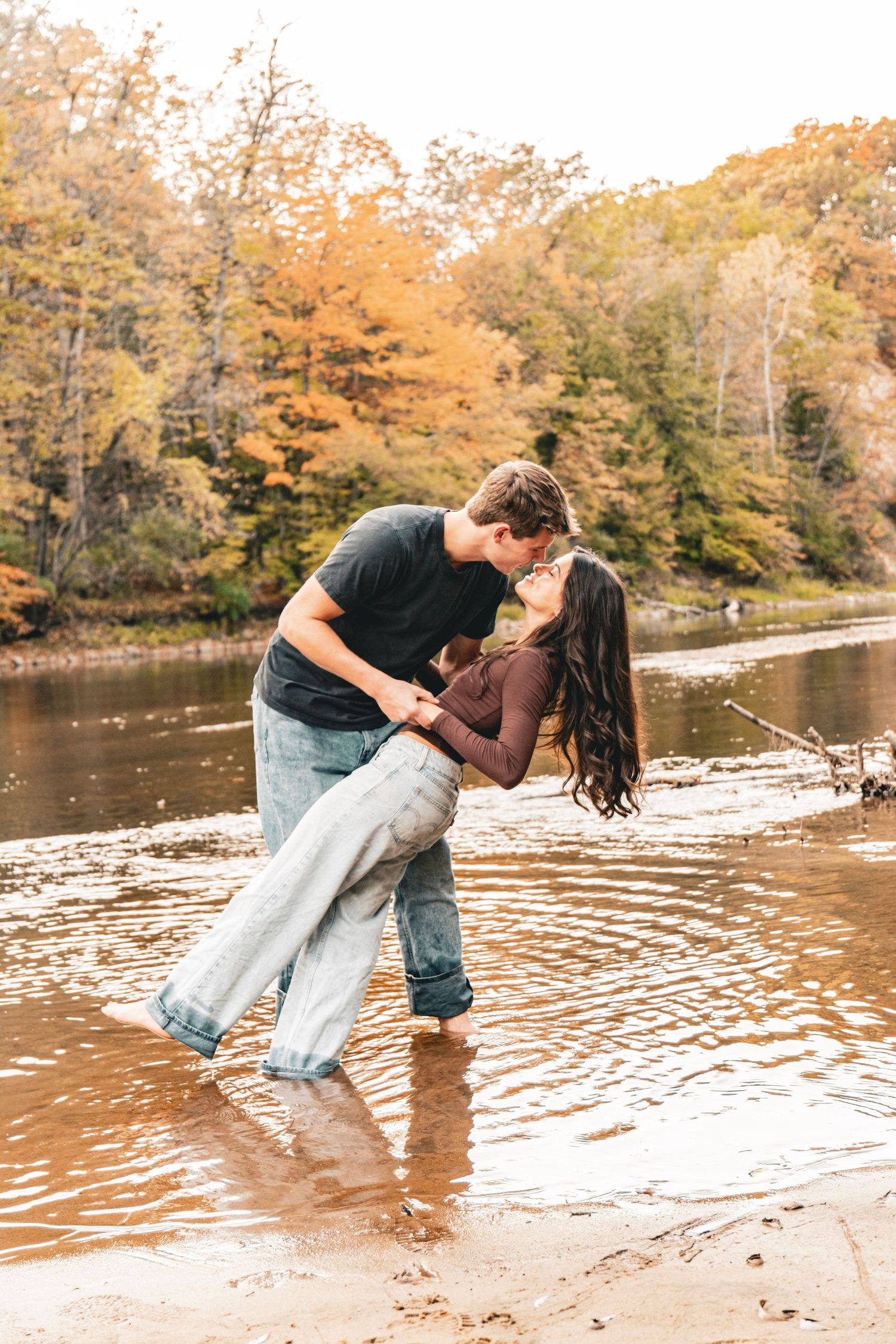 Couple in casual clothes share a romantic dip kiss while standing in shallow river water, with colorful autumn trees along the shoreline.