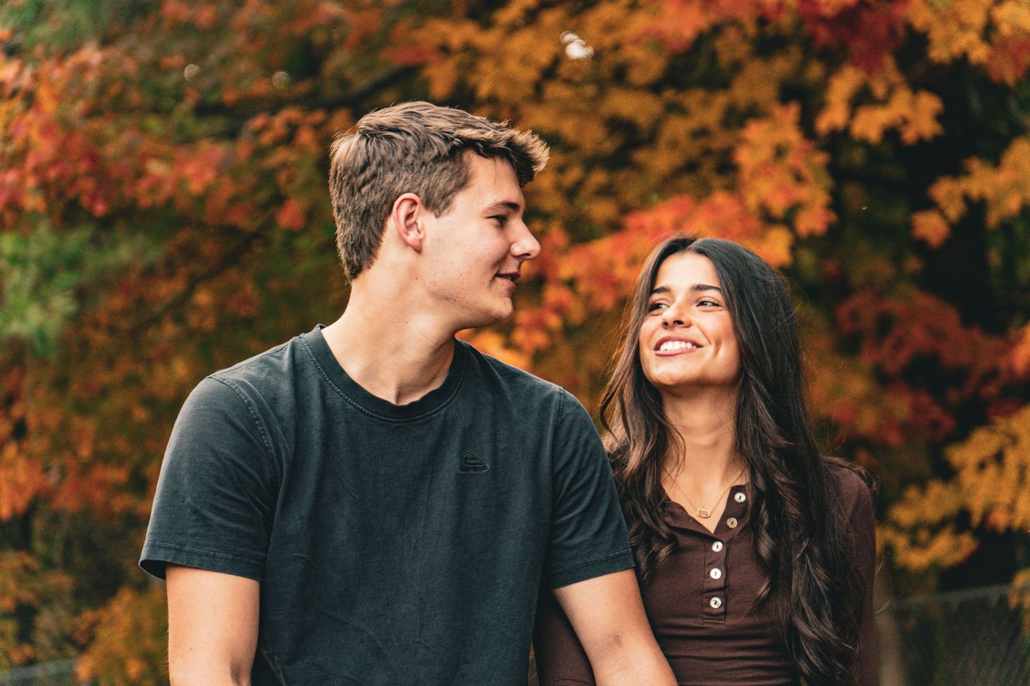 Smiling young man and woman sitting close together outdoors, enjoying a happy moment against a backdrop of colorful autumn foliage.