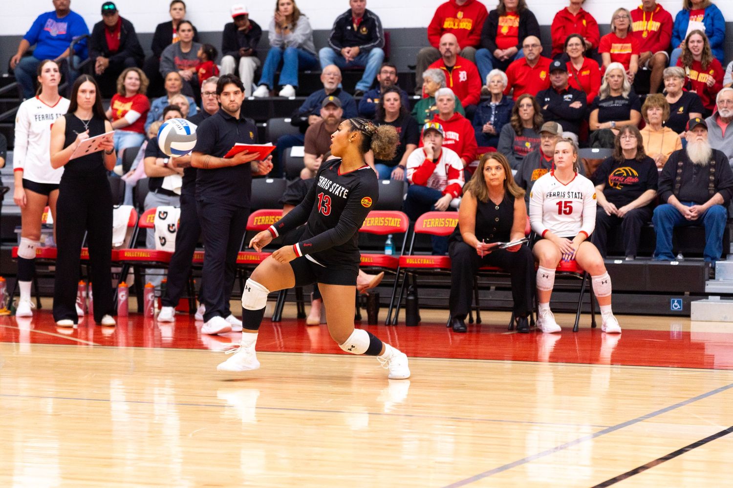 Volleyball player in a black jersey crouches in a ready stance on the court as teammates and a crowd in red watch from the sideline.