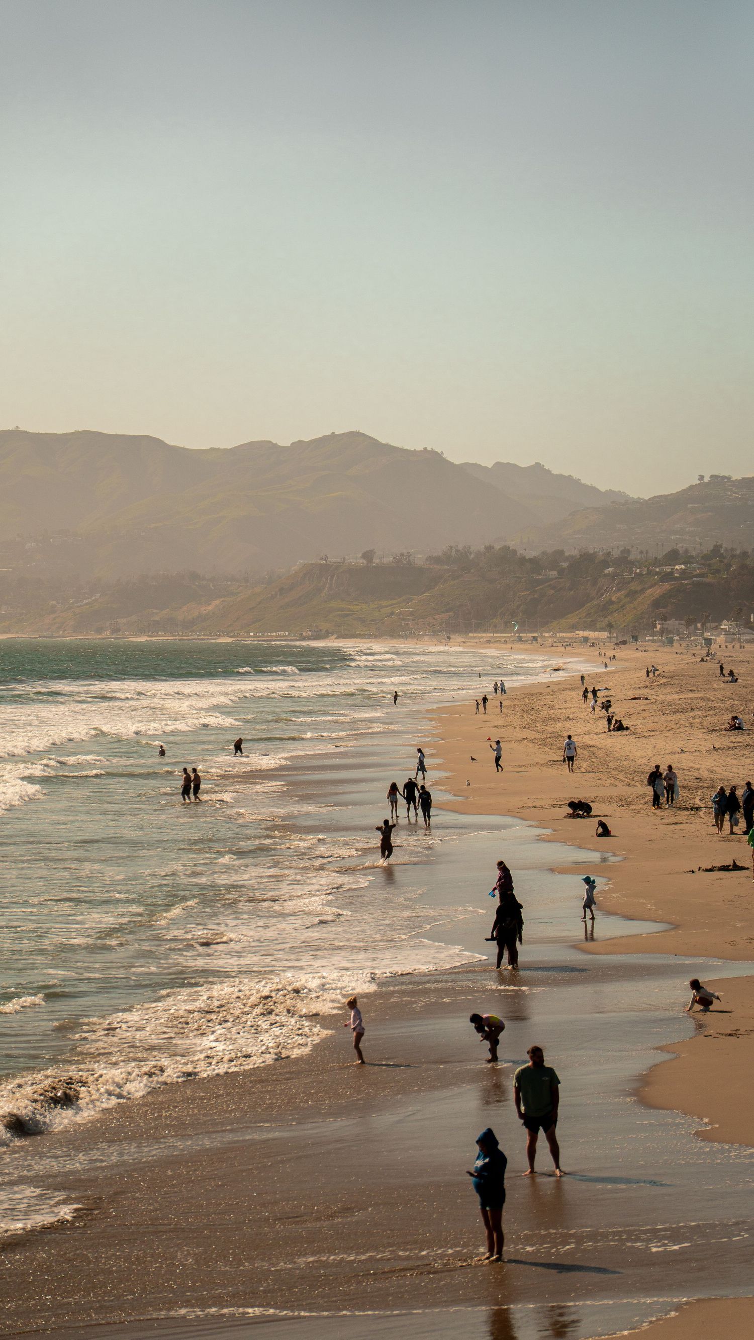 Beachgoers strolling along the shoreline as waves crash on a cloudy afternoon.