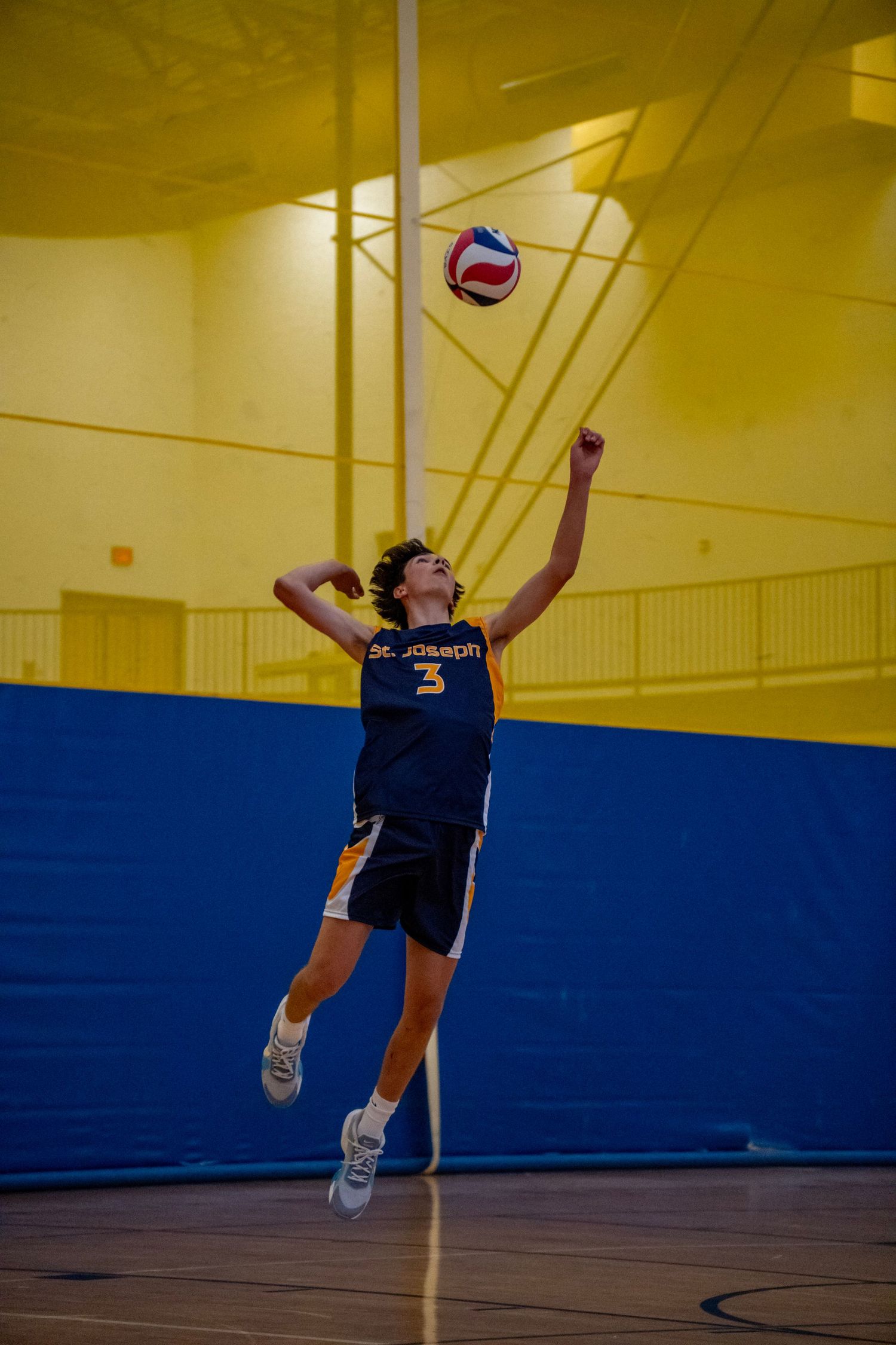Volleyball player in navy uniform jumps to serve the ball in a yellow-lit gymnasium.