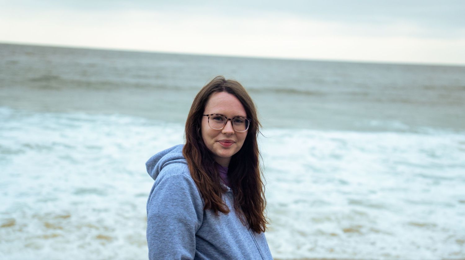 Someone in casual beachwear strolls along the sand as waves roll in on a cloudy day.