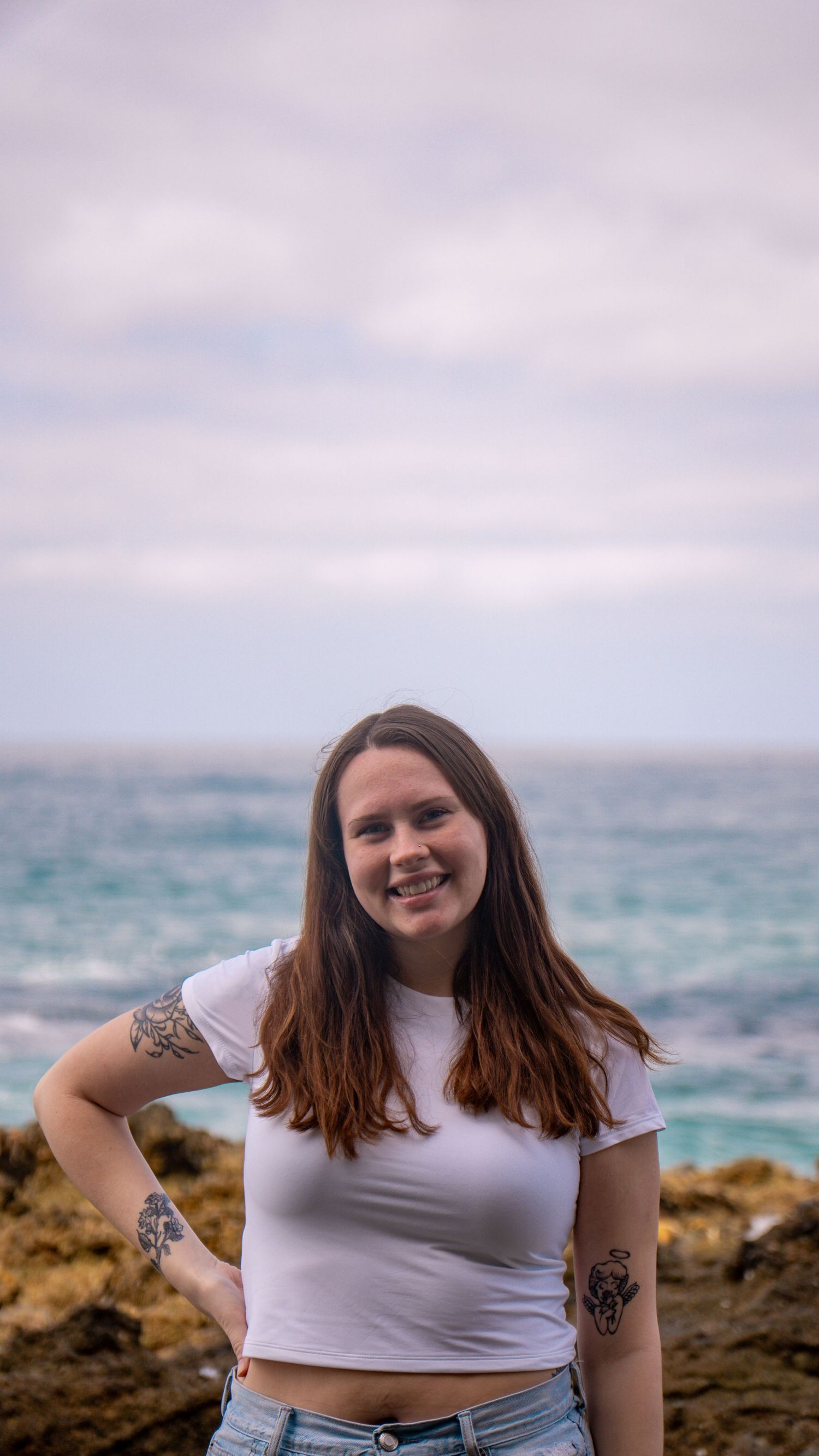 Beachside portrait with visible tattoos against cloudy coastal backdrop.