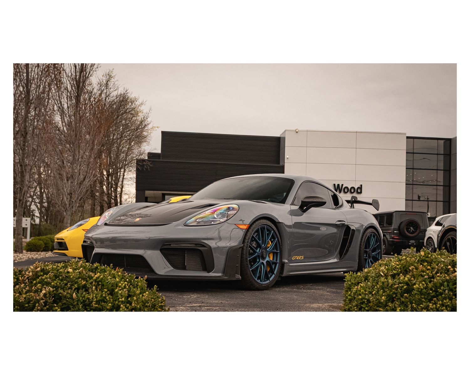 Gray Porsche GT car with blue rims parked in front of modern building with yellow car in background.