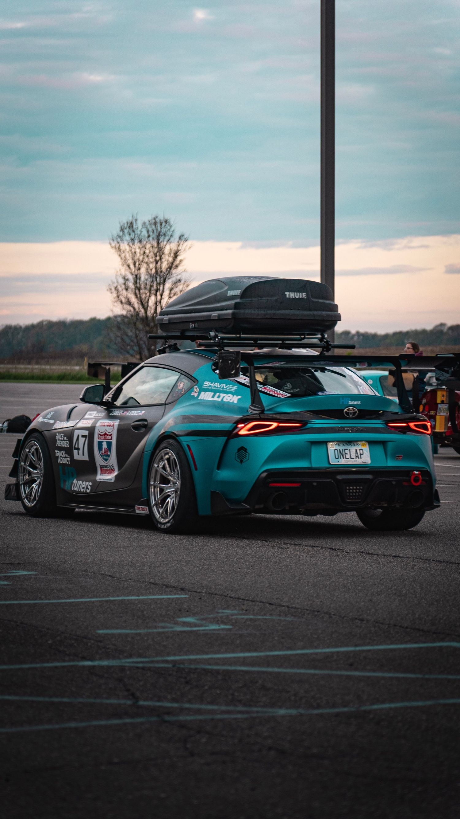 Teal race car with roof cargo box parked at sunset with dramatic sky in background.