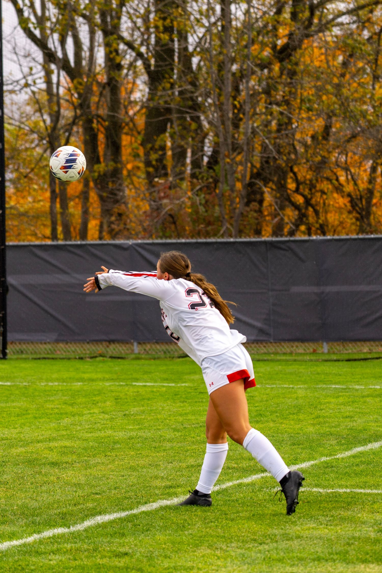 Soccer player in white uniform throws ball from sideline during autumn match on grass field with fall foliage in background.
