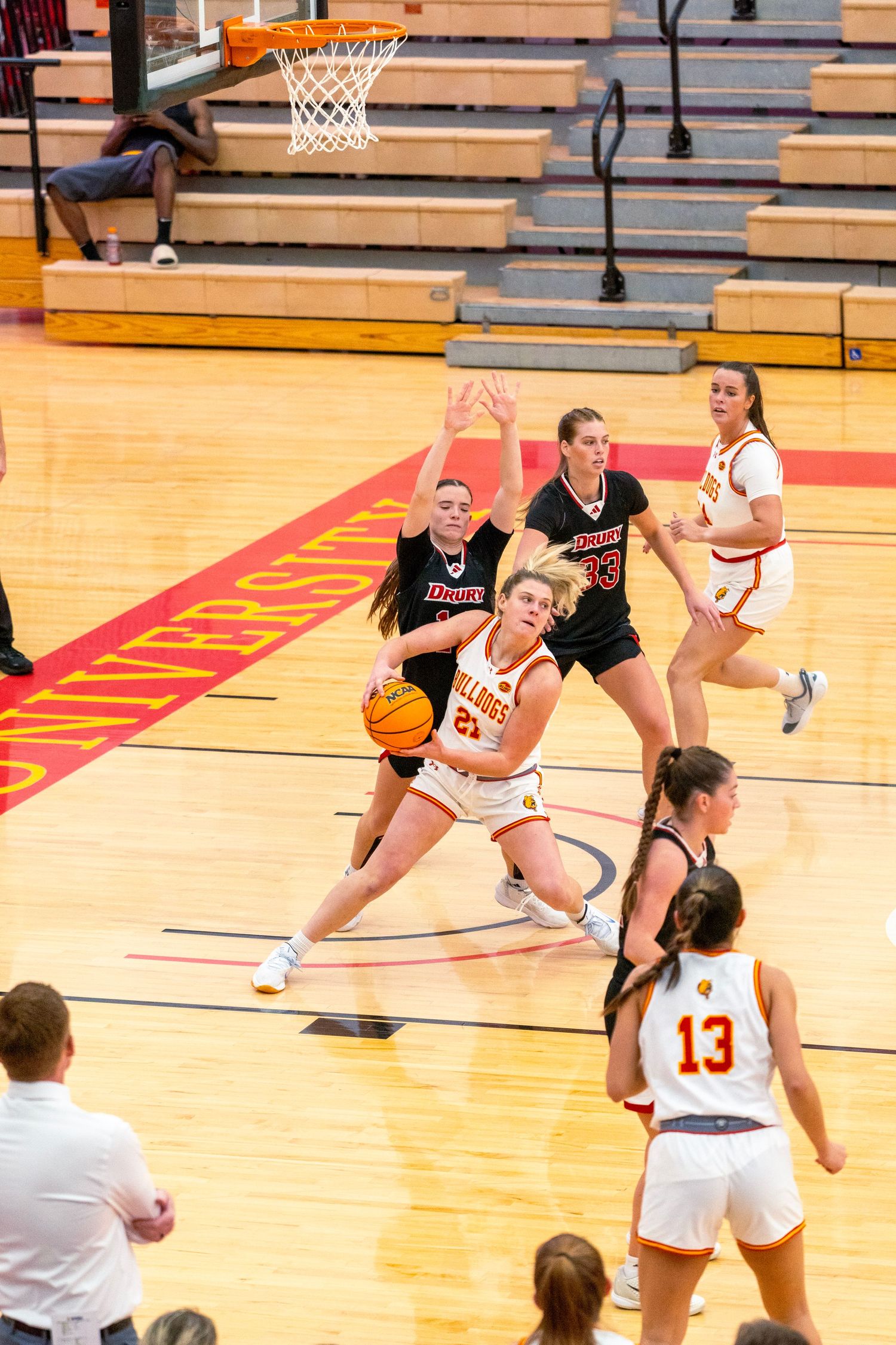 Female basketball players compete on a hardwood court during an intense game with spectators in the background.