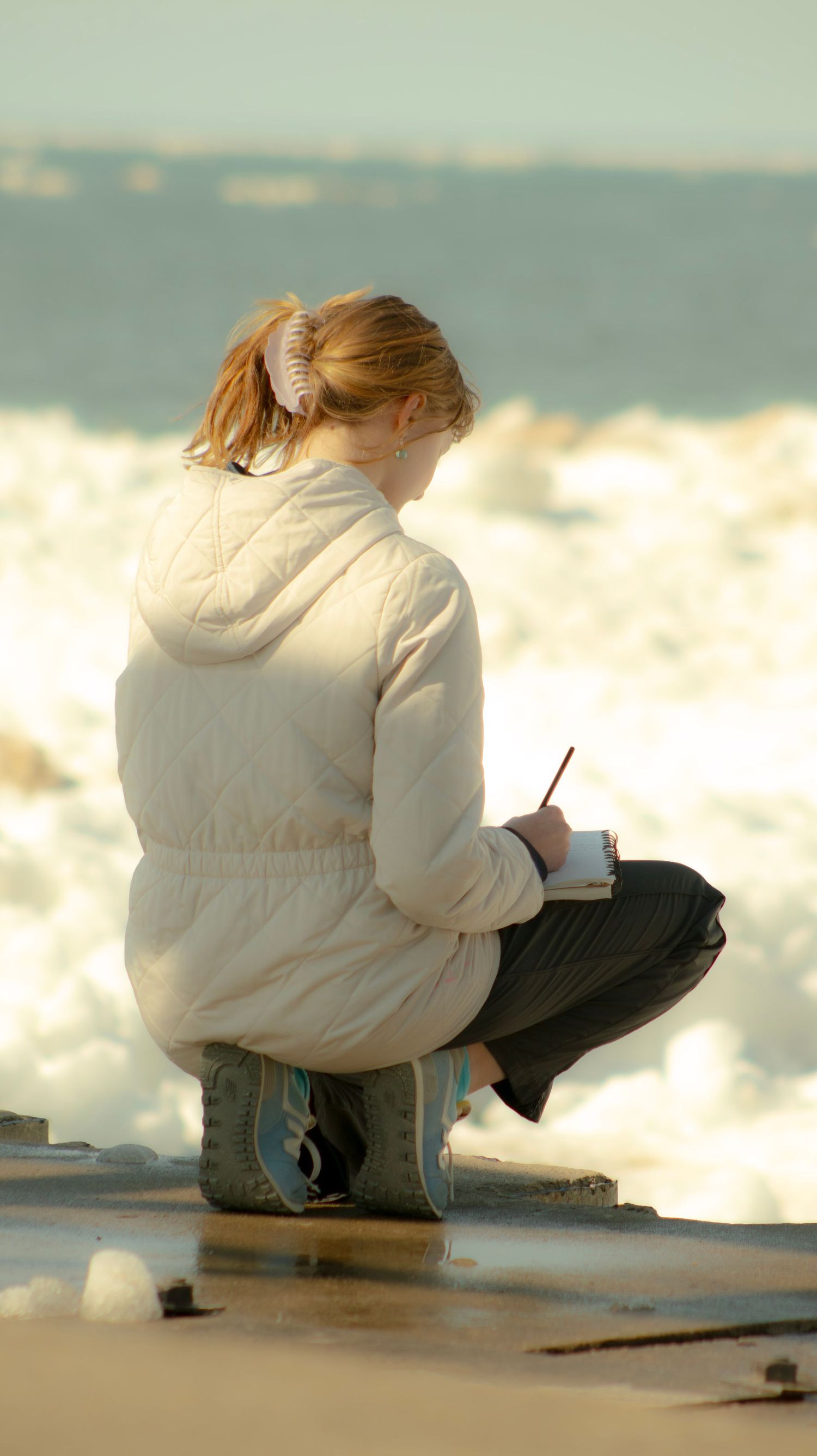 Person sitting by the ocean at sunset wearing a white sweater and capturing the peaceful view.