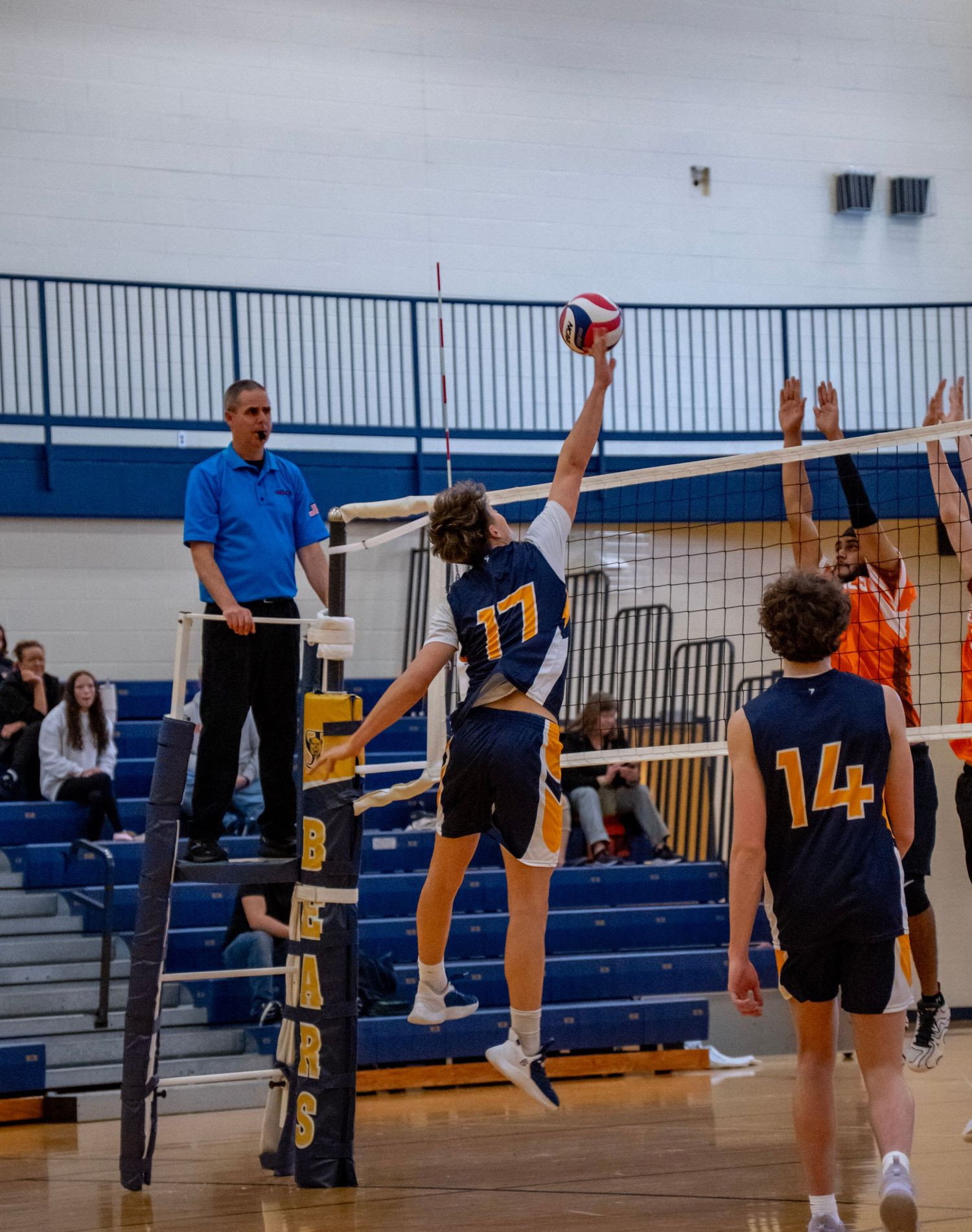 Teams face off at the volleyball net with players jumping to block a spike.