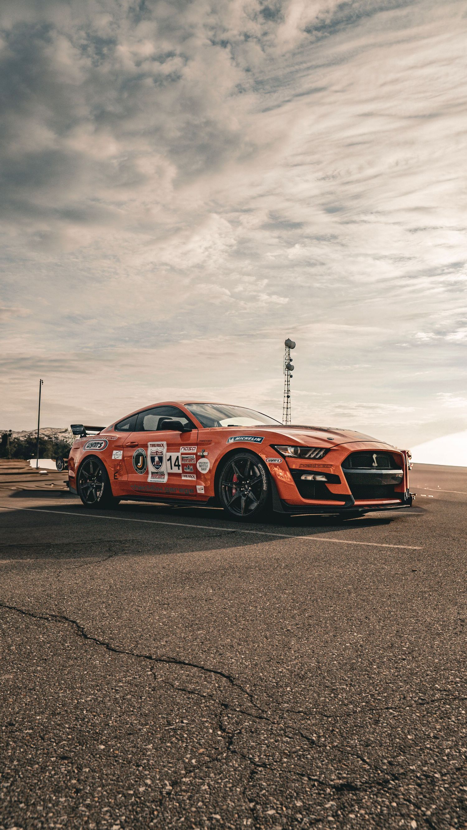 Orange racing Mustang Shelby GT500 with competition livery parked on asphalt under dramatic clouds.