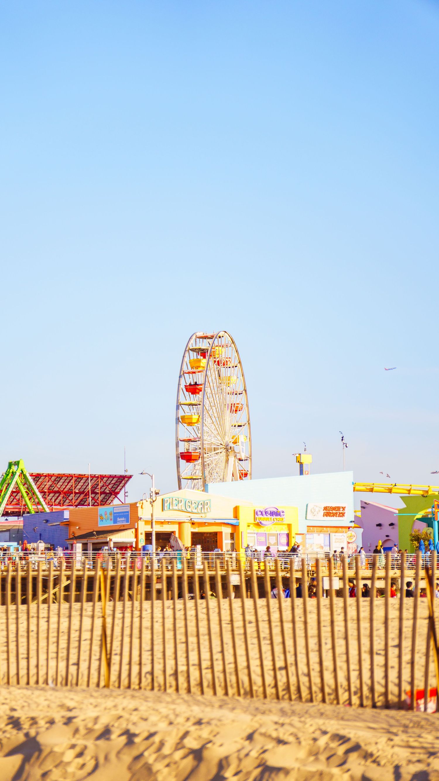 Wooden fence posts line a beach boardwalk with colorful amusement park rides visible in the distance.