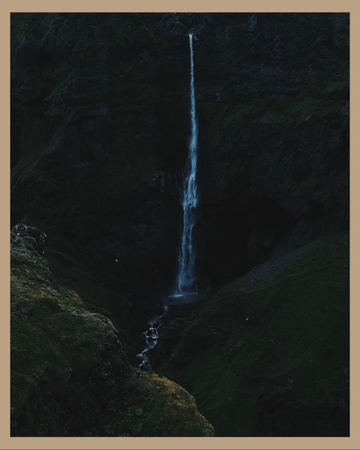 Thin waterfall cascading down dark rocky cliff face in dramatic natural setting.