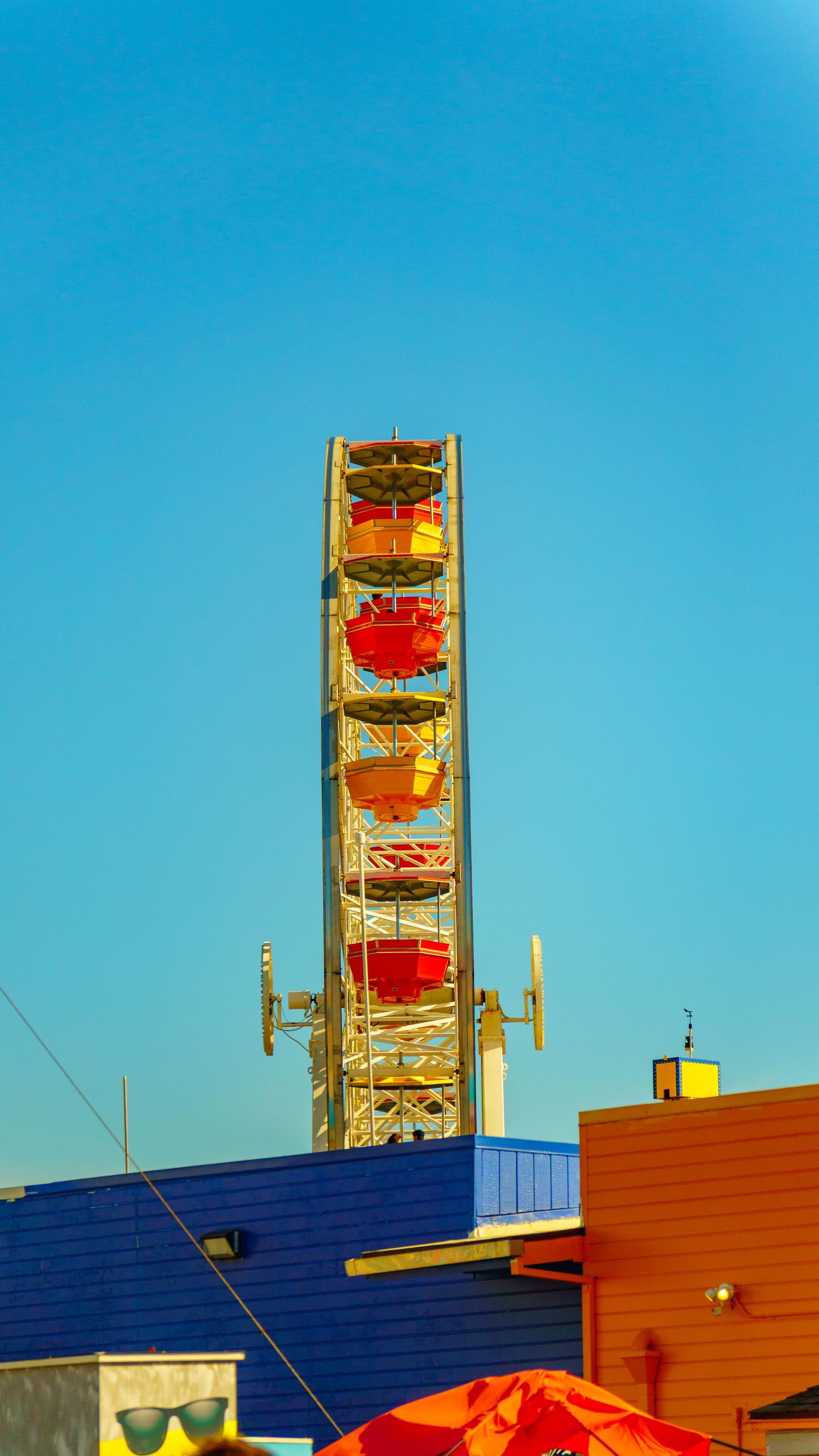 Colorful Ferris wheel cars against bright blue sky with geometric buildings below.