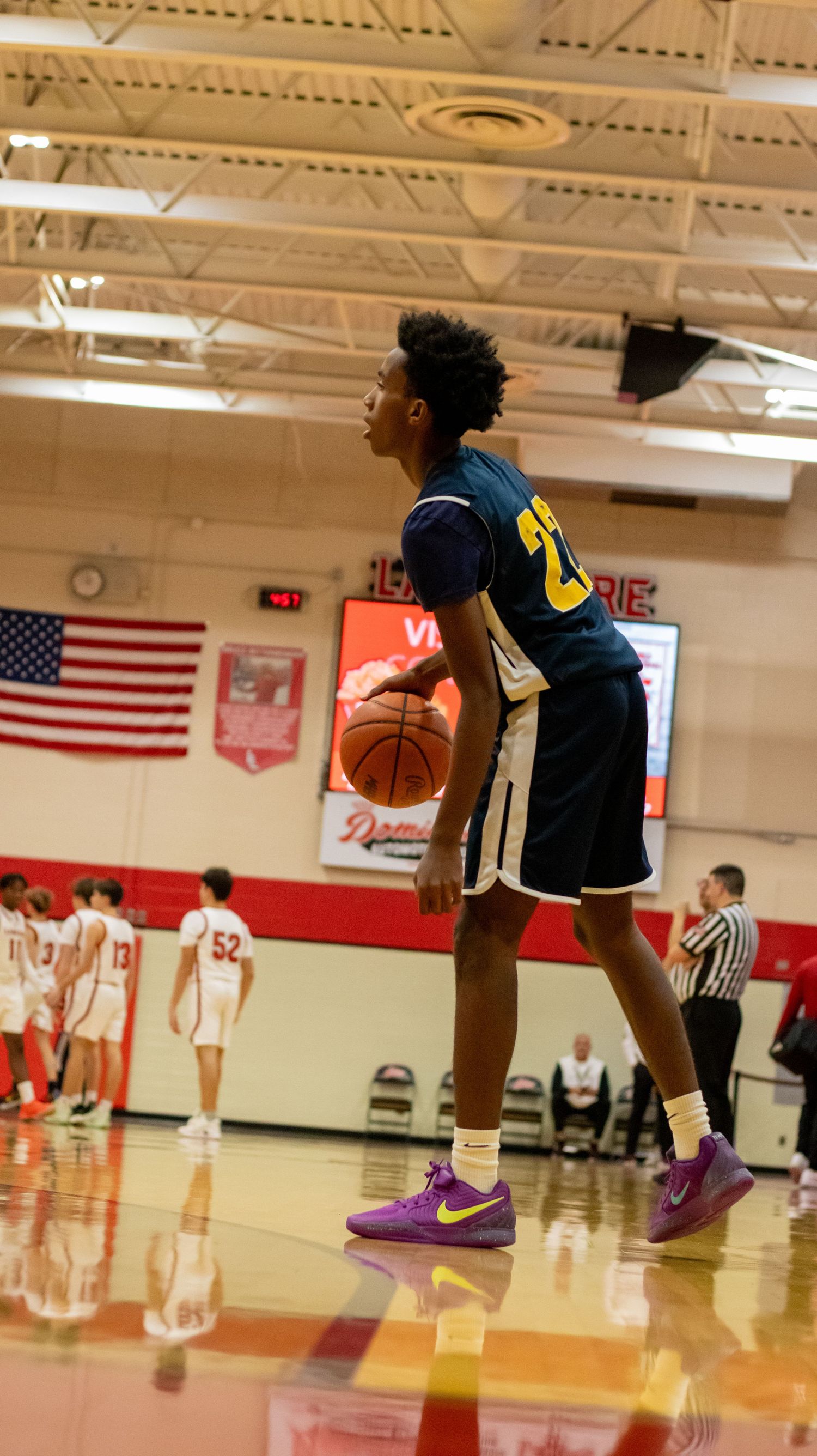 Players in navy blue and white uniforms position themselves during a basketball game with American flags visible.