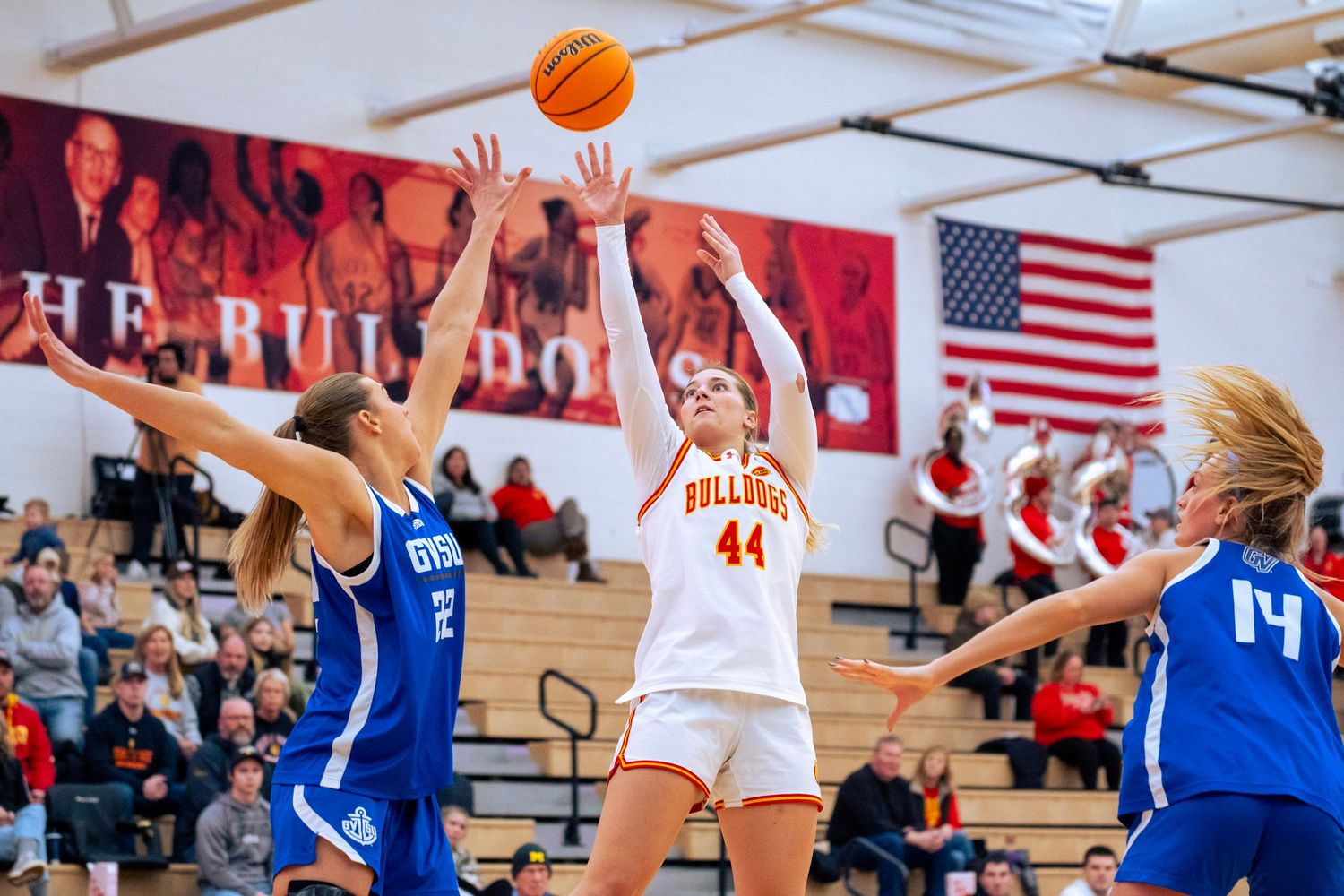 Women's basketball player in a white number 44 jersey shoots over two defenders in blue uniforms during an indoor game with fans watching.