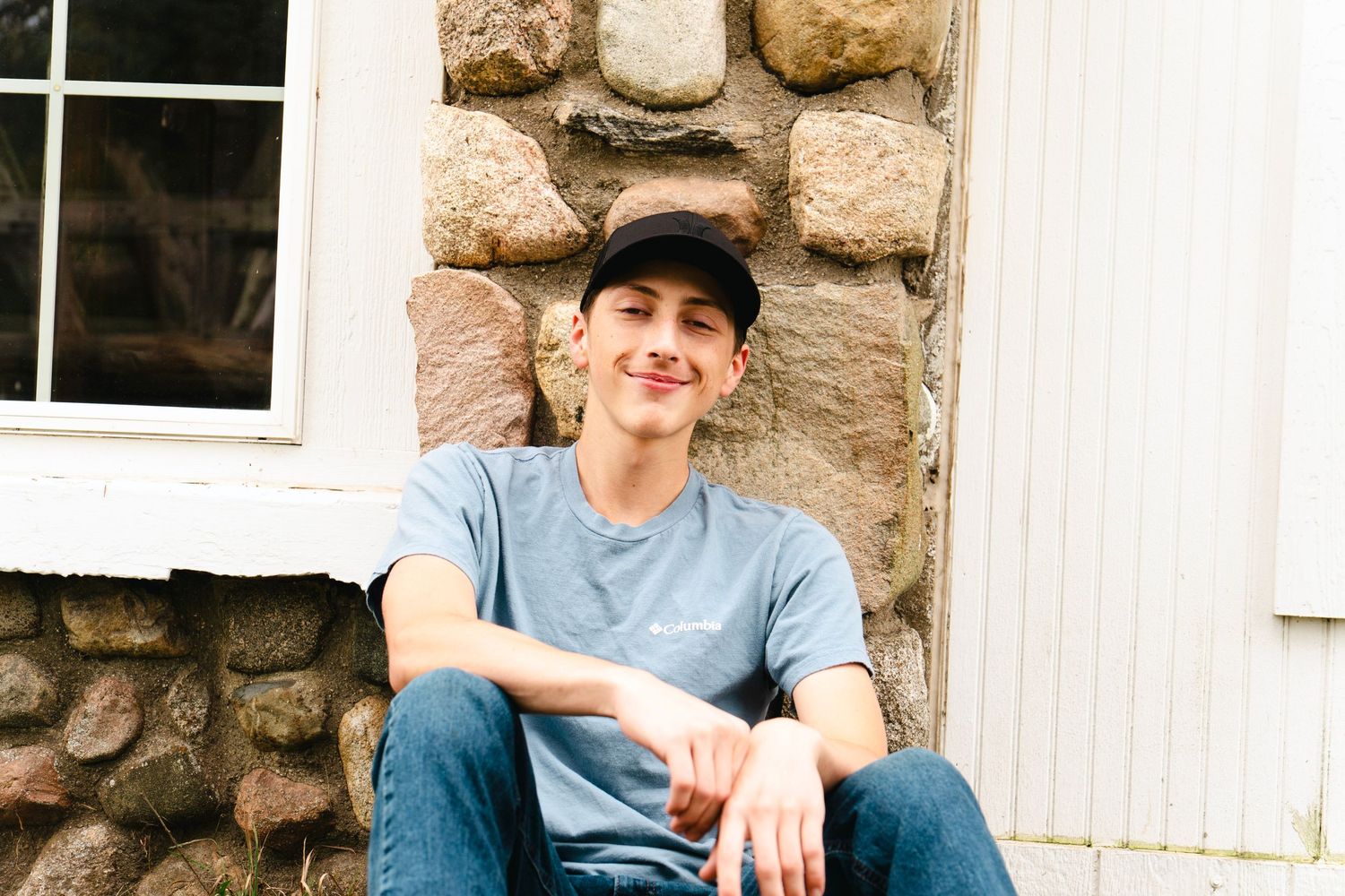 Smiling young man in a black cap, gray t-shirt and jeans sitting on stone steps beside a rustic stone wall and white siding.