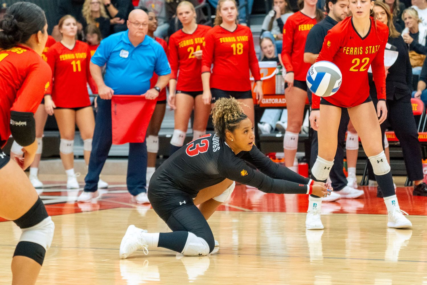 Volleyball players stretch and prepare on a glossy indoor court with team banners visible in the background.