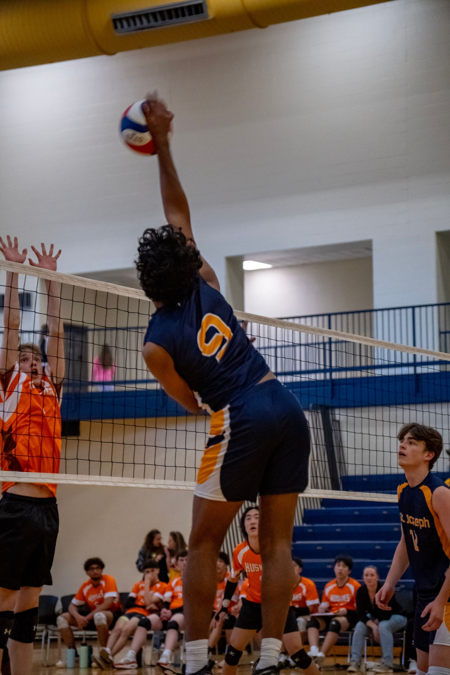 Player in navy jersey spikes volleyball against orange-clad defenders at the net.