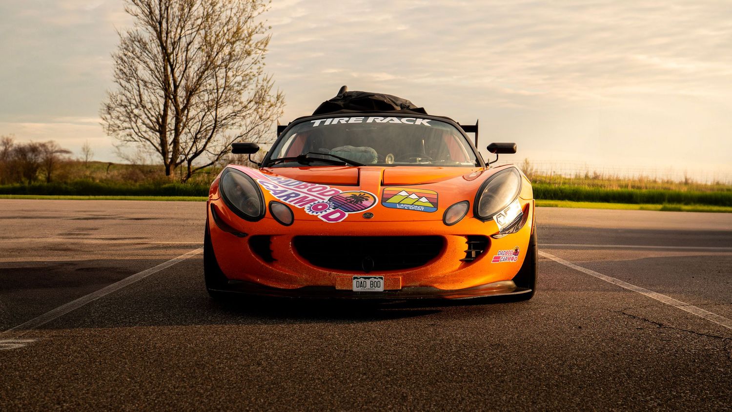 Front view of orange Lotus race car with custom livery parked on asphalt.
