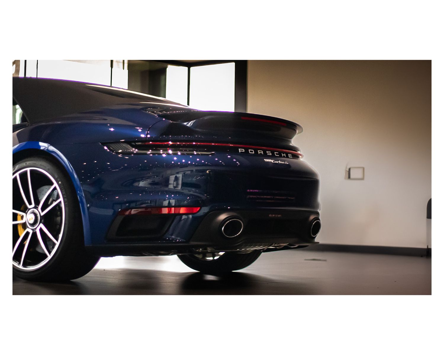 Close-up view of a dark blue Porsche sports car rear end with dual exhaust and spoiler in a garage setting.