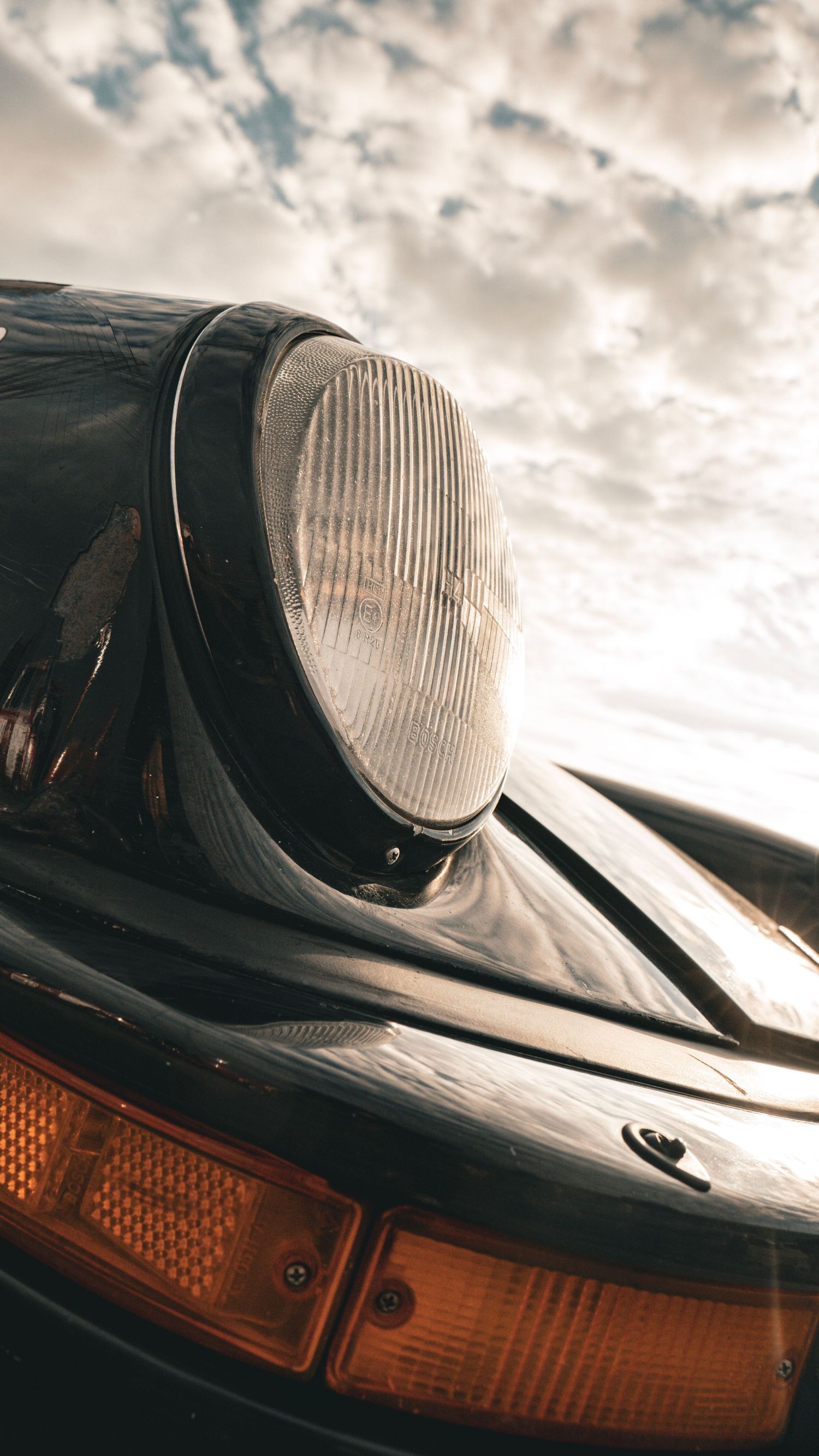 Close-up detail shot of a classic Porsche 911 headlight and front bumper against cloudy sky.