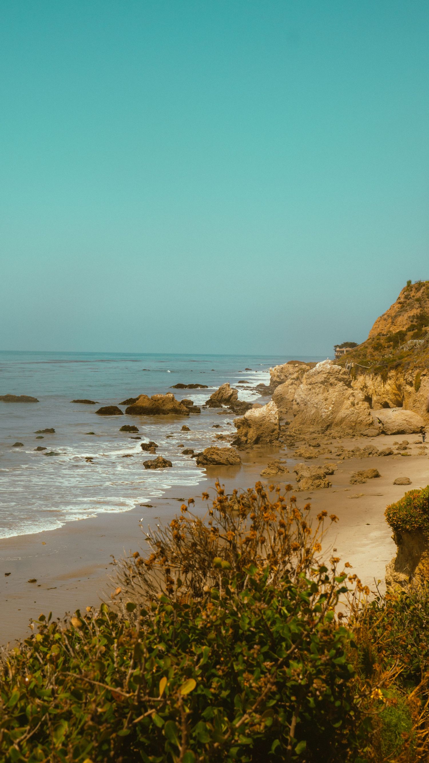 Rocky coastal cliffs and sandy beach meet the turquoise ocean waters under a calm blue sky.