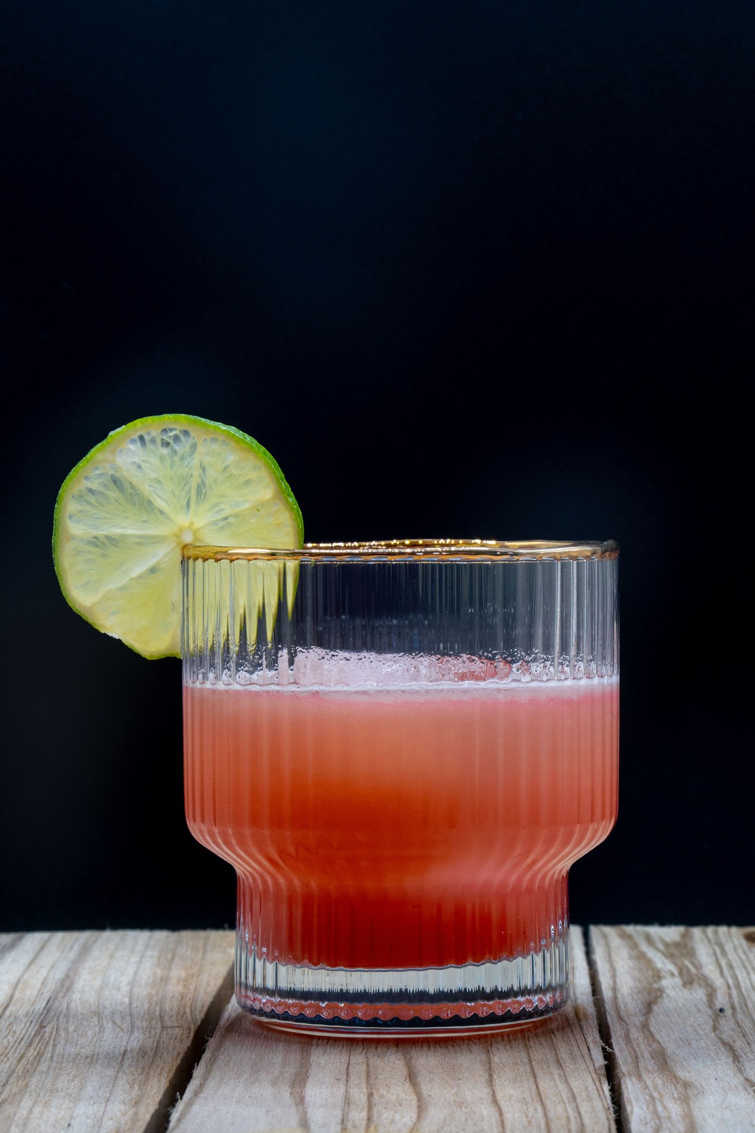 A red cocktail with lime garnish in a vintage-style coupe glass on a rustic wooden surface.