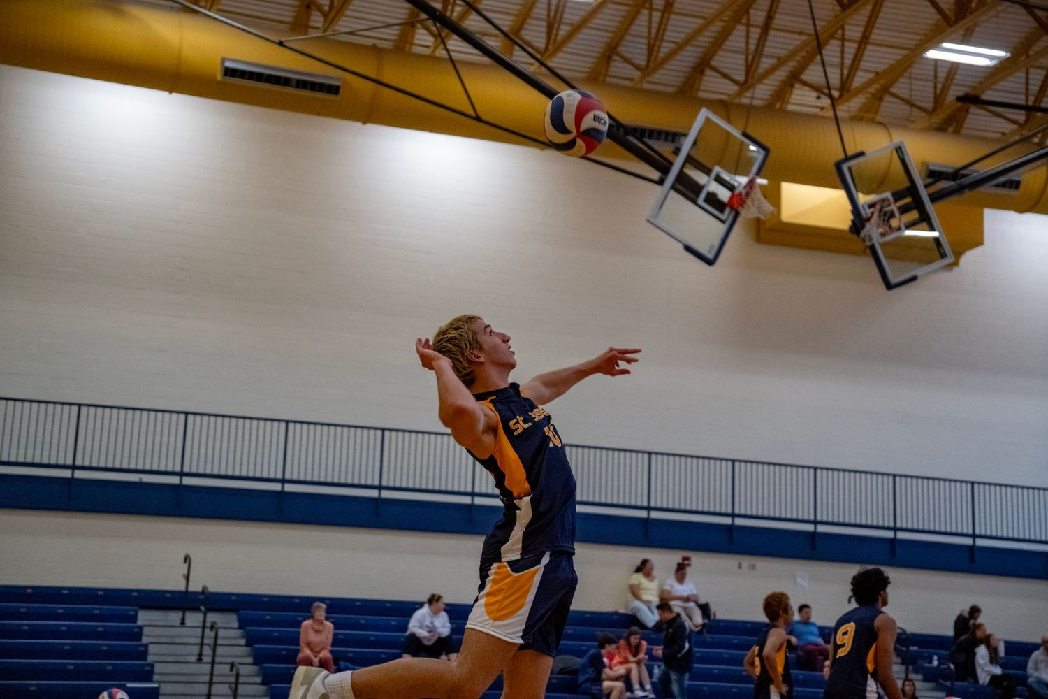 Volleyball player jumps high to execute a serve in a gymnasium with yellow lighting.