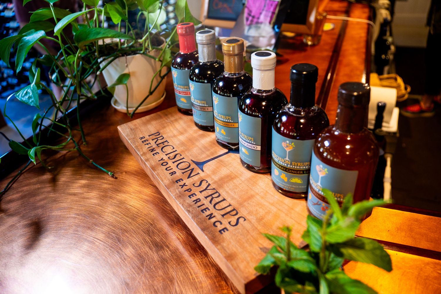 Row of specialty cocktail syrups displayed on wooden board with decorative plants.