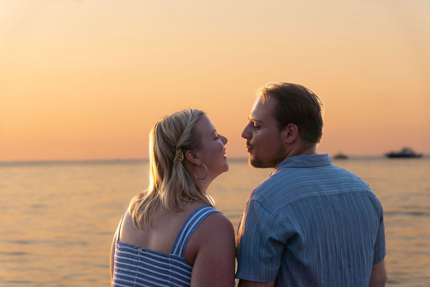 Couple shares intimate moment at sunset by ocean with warm orange sky and calm waters in background.