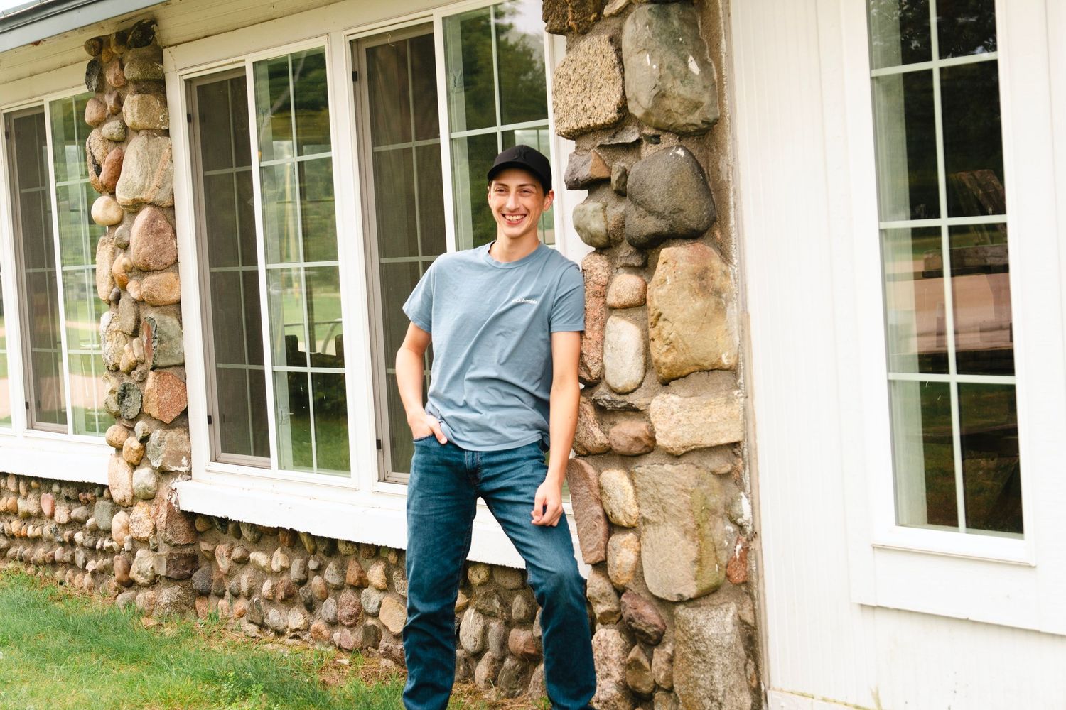 Young man in a gray t-shirt, jeans and cap leans against a fieldstone wall beside large windows of a white house with green grass.