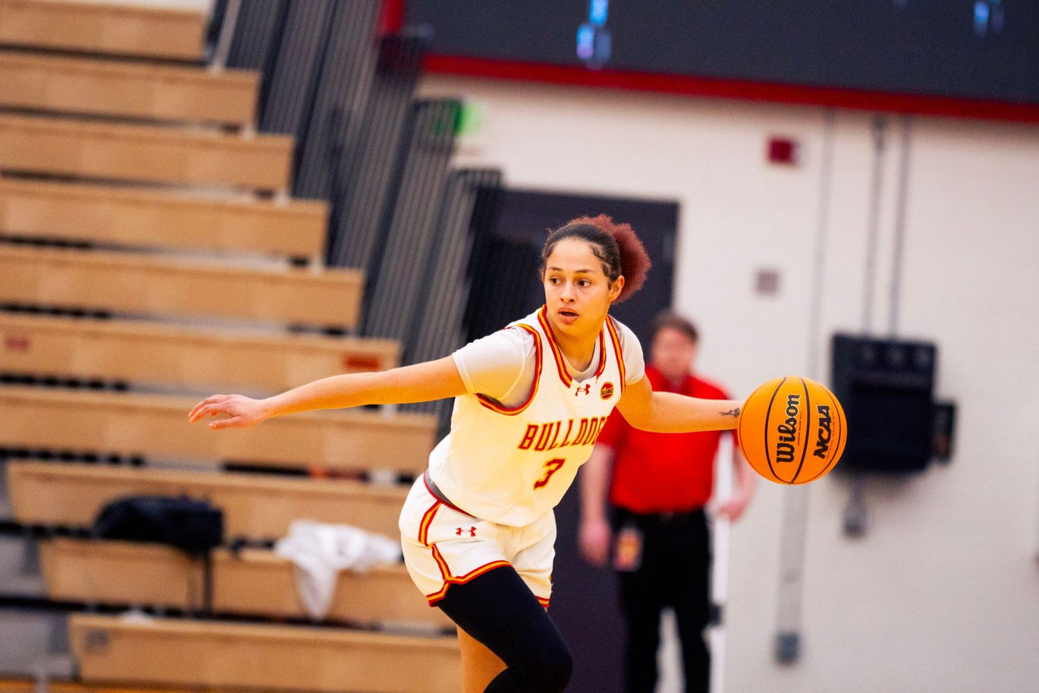 Female Bulldogs basketball player in a white jersey dribbles the ball on the court with empty wooden bleachers and a referee behind her.