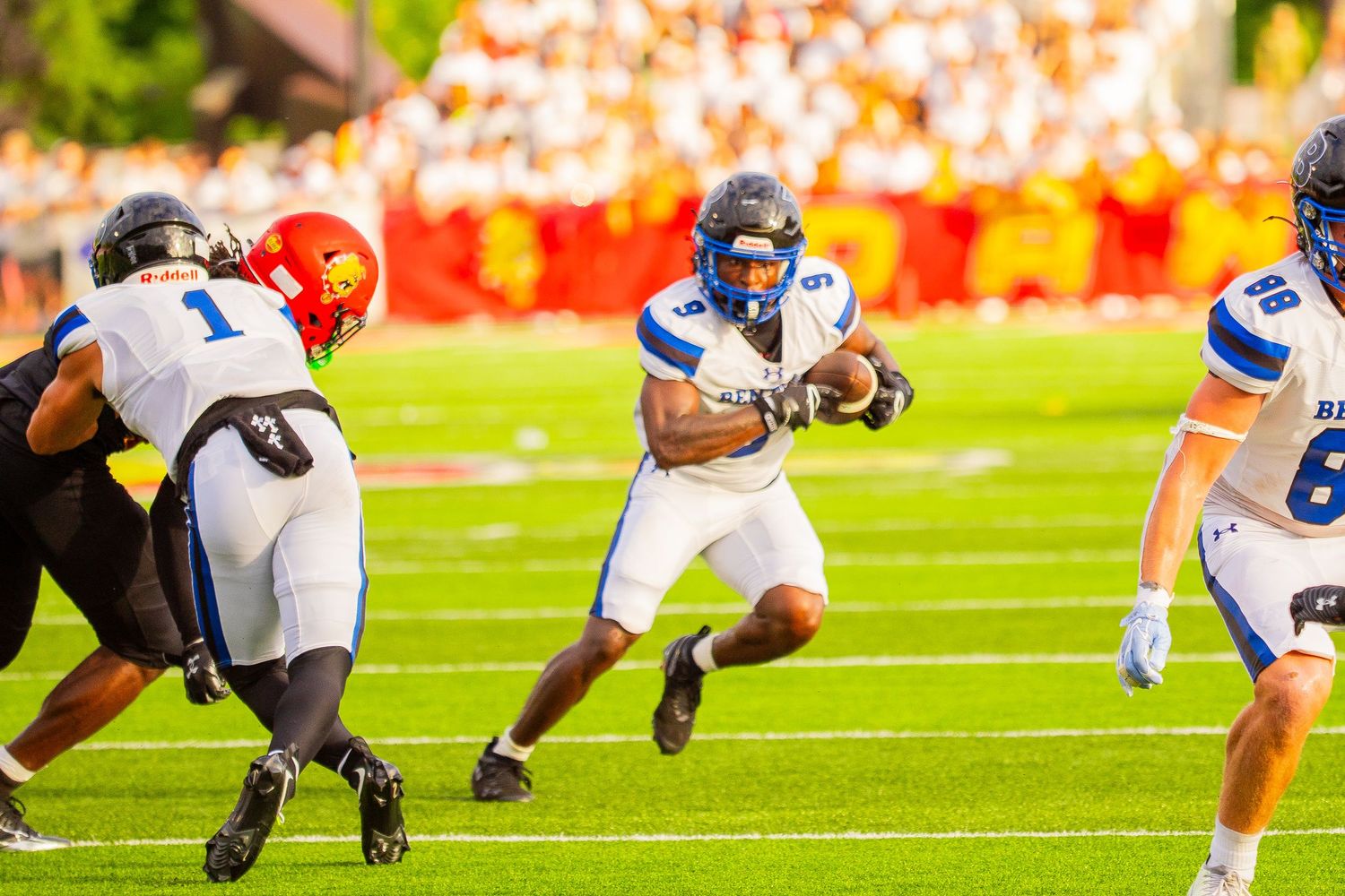 A football running back in a white and blue uniform carries the ball upfield as defenders in white and red chase him during a game.