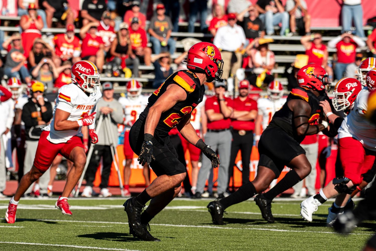 Football players in red and white uniforms compete during a sunny game with fans watching from the stands.