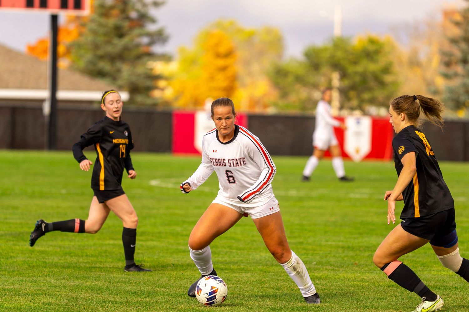 Soccer players compete for ball possession during an intense match on a grass field in autumn weather.