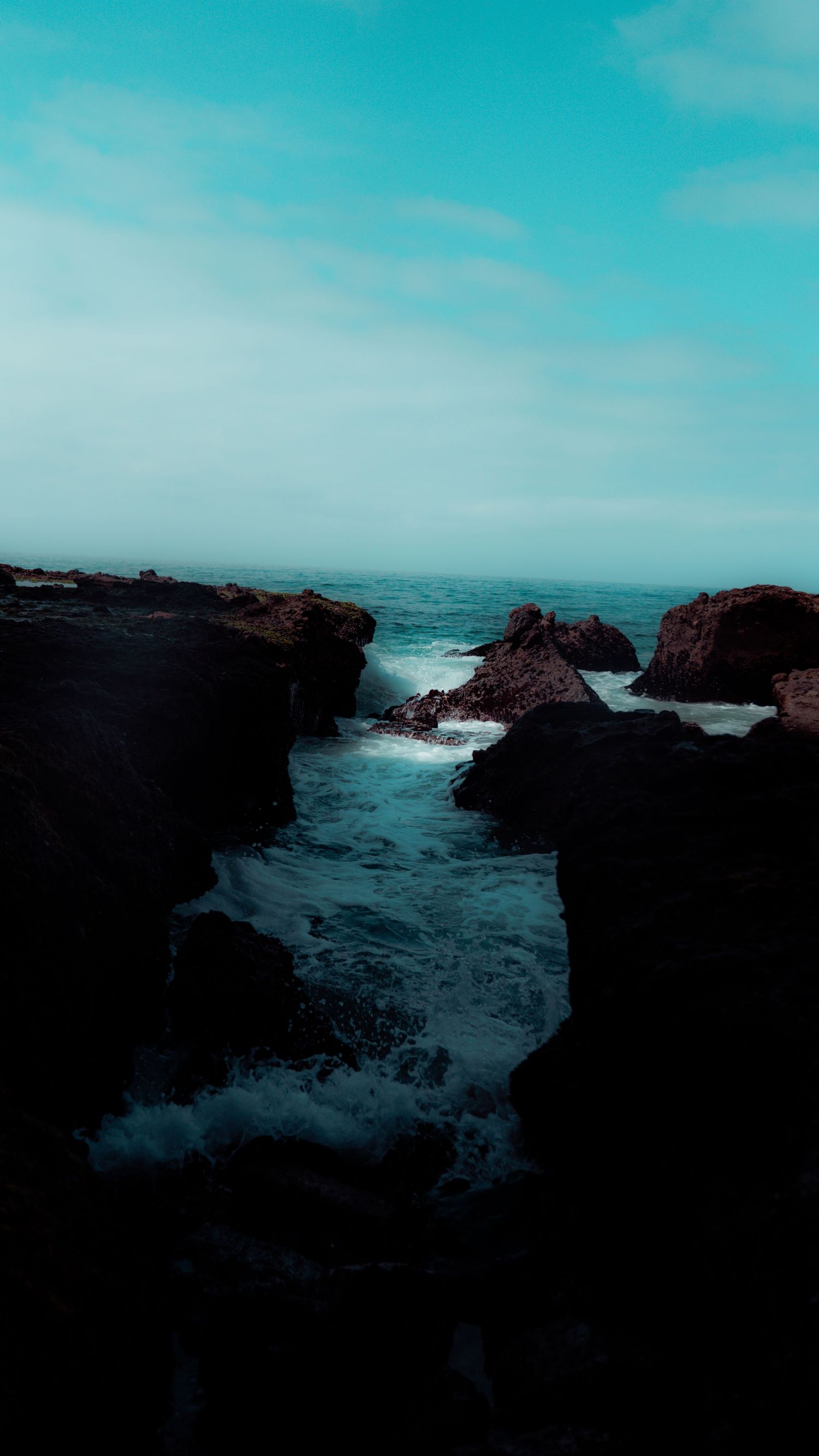 Dark rocky cliffs frame a narrow ocean channel with crashing waves.