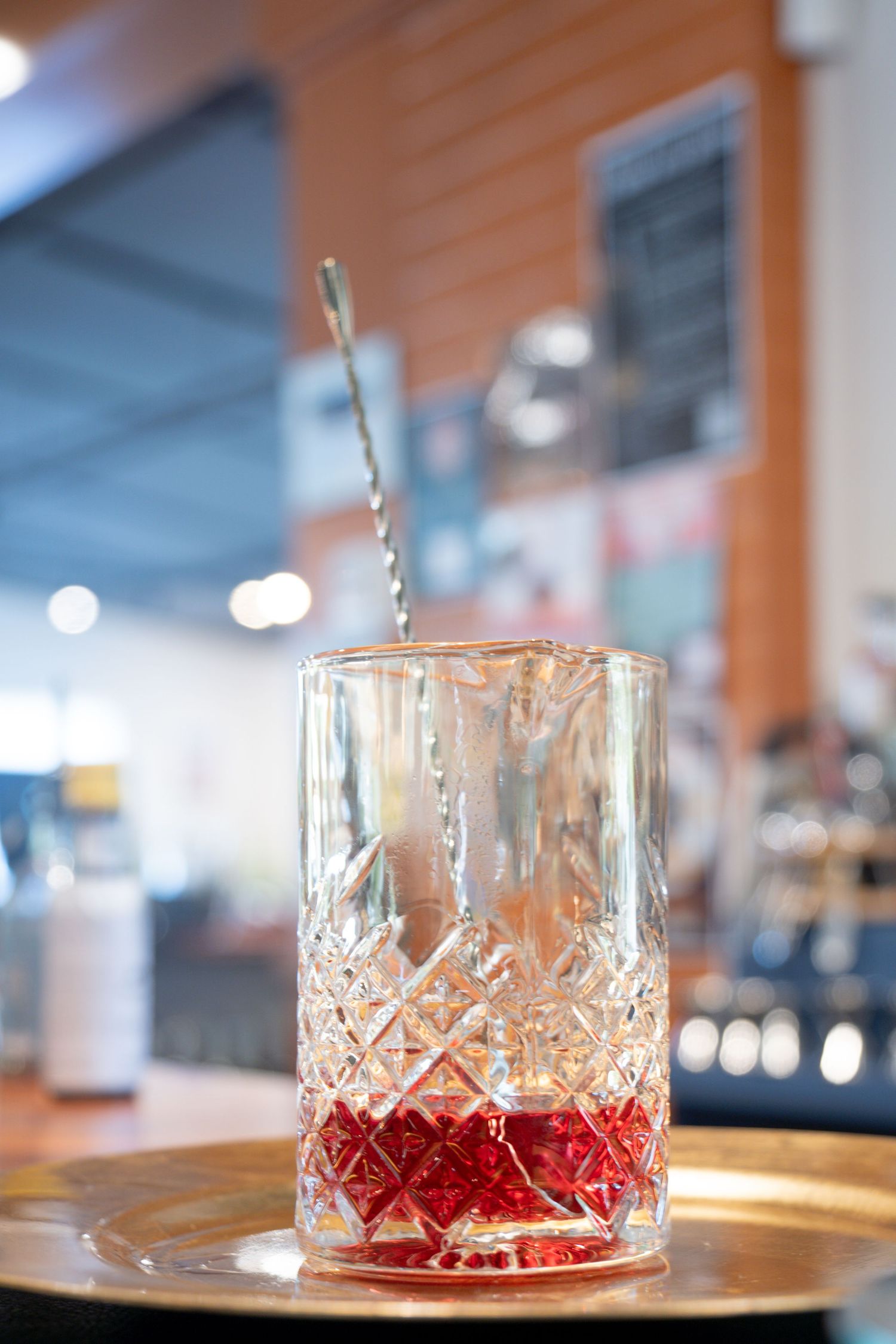 Crystal cocktail glass with red syrup and mixing spoon on wooden surface in bar setting.