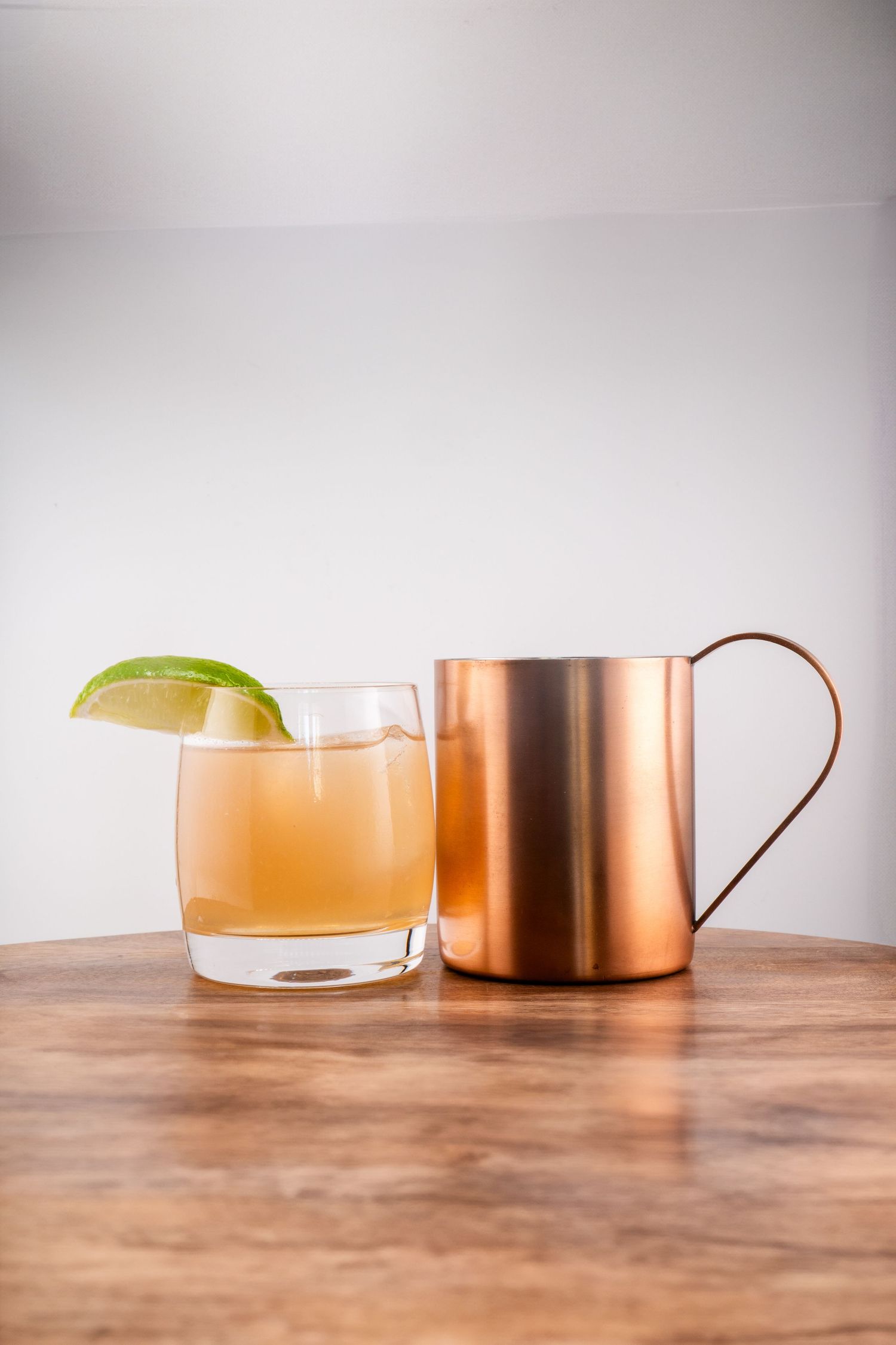 Classic cocktail served with lime garnish beside copper mug on wooden table.