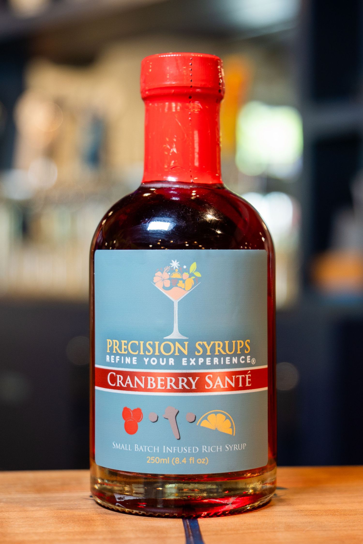 A bottle of Precision Syrups Cranberry Sante cocktail mixer with a blue label and red cap on a wooden surface.