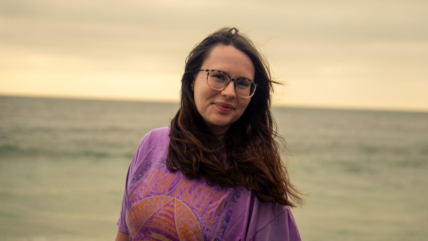 A person in casual attire enjoys a peaceful moment at the beach with waves and cloudy skies in the background.