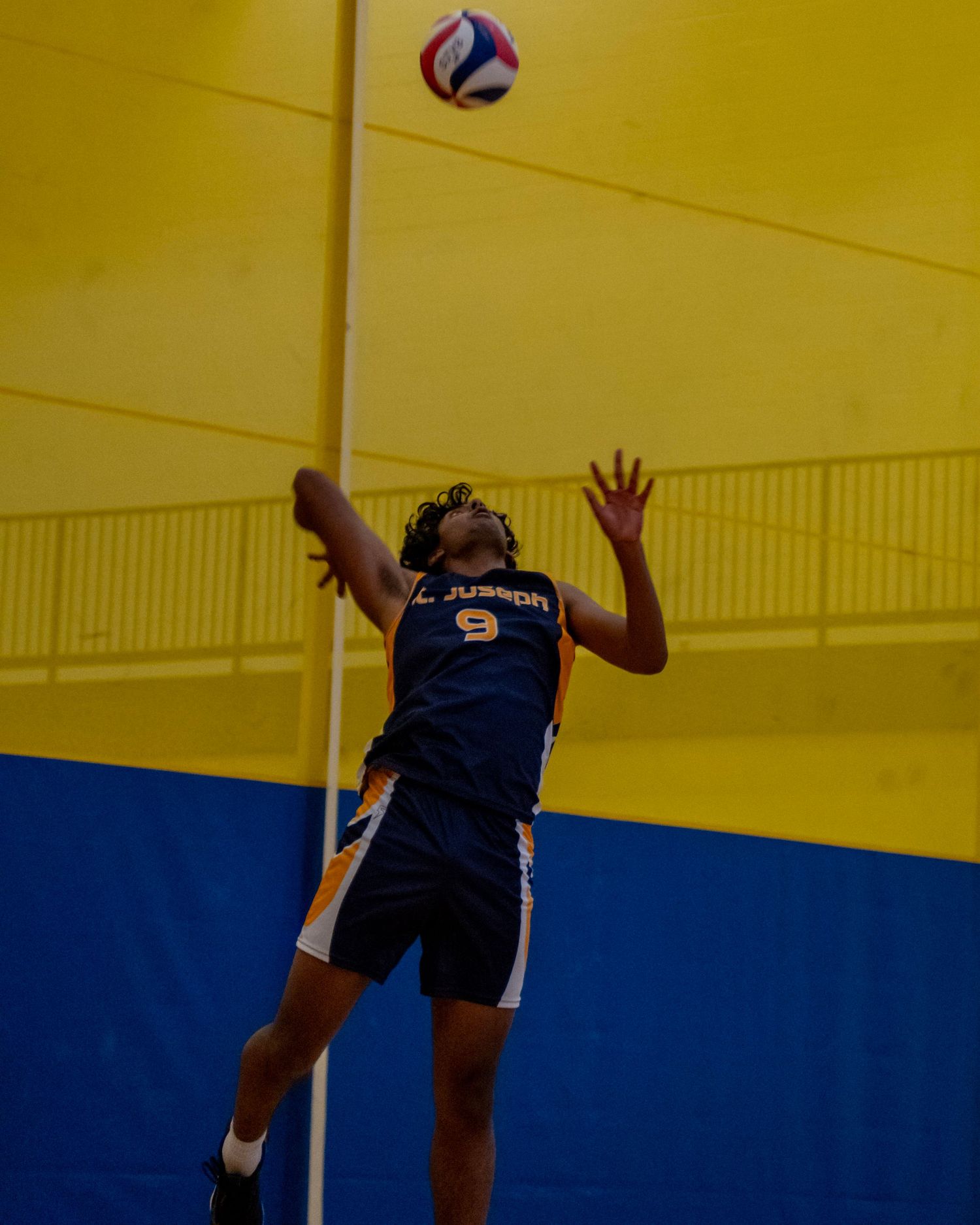 Player jumping to spike volleyball in navy uniform against yellow gymnasium wall.