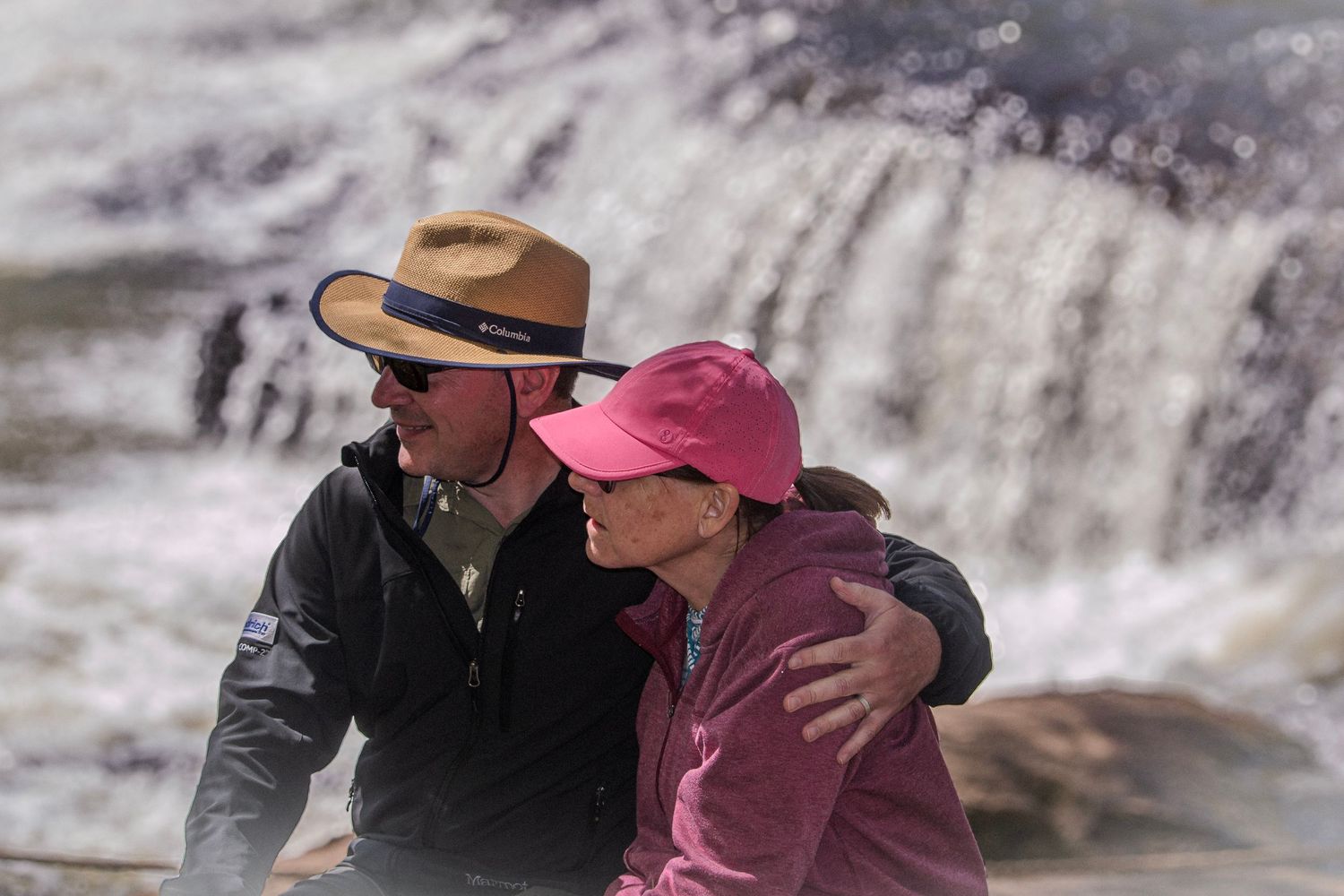 Two people embrace while watching a waterfall in nature, one wearing a brown hat and the other in a pink jacket.