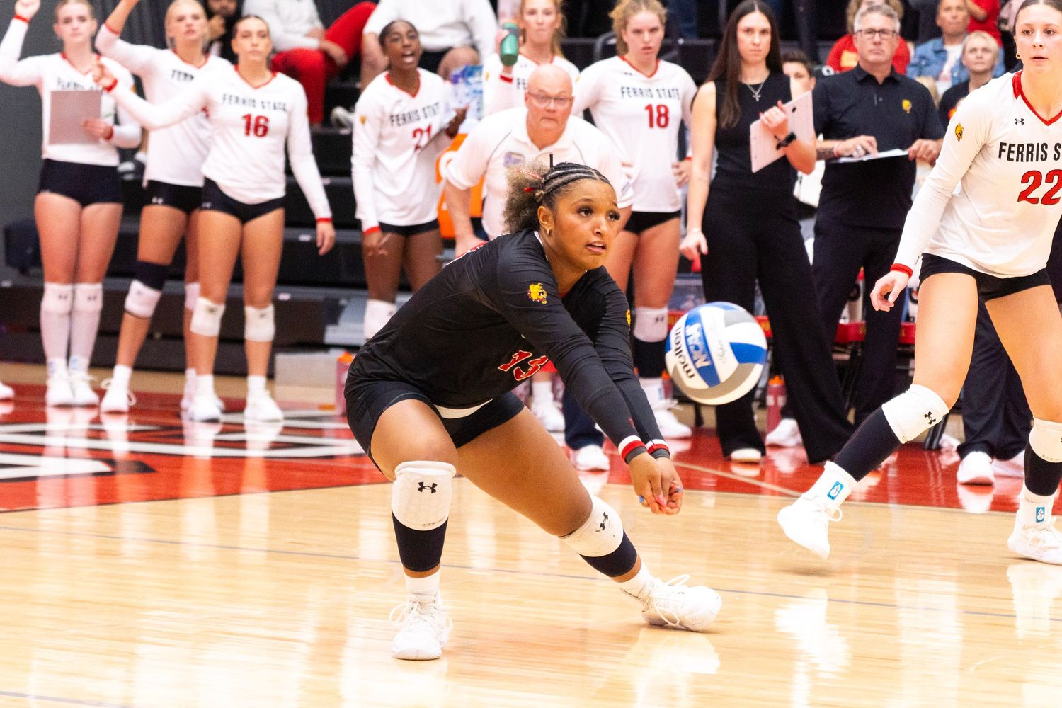 A volleyball player in a black uniform digs a low pass on a hardwood court while teammates in white jerseys watch from the sideline.