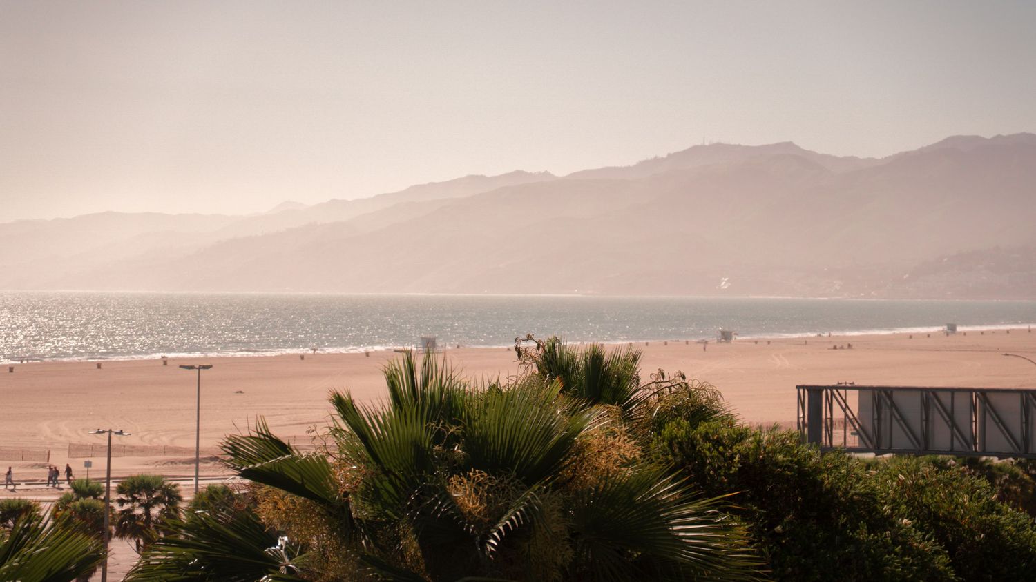 Coastal landscape with palm trees and mountains overlooking a sandy beach at sunset.