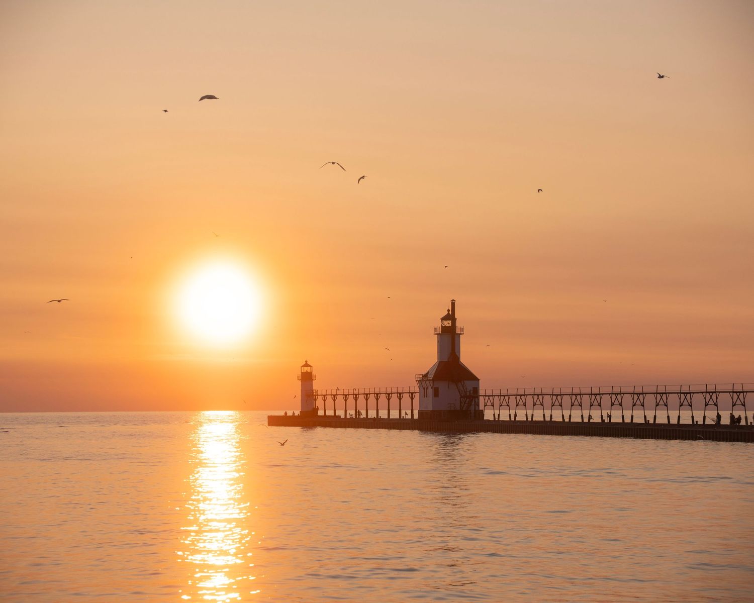 Historic lighthouse and pier extending into calm waters during golden sunset with seabirds flying overhead.